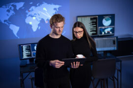 Junger Mann und junge Frau vor Monitoren schauen auf ein Laptop
